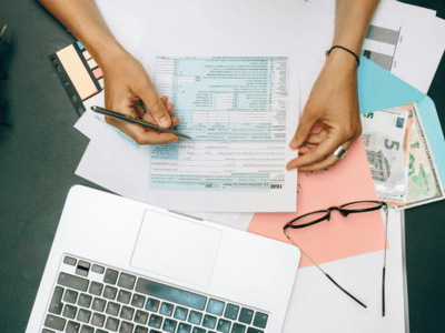 A person filling forms in front of a laptop with glasses and some currency notes on the surface.