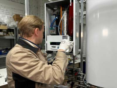 Technician repairing a residential heating system with an open access panel showing internal pipes and controls.